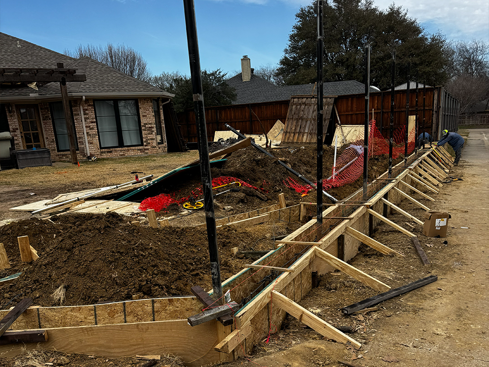 Backyard construction site with wooden trench forms, dirt piles, and black vertical pipes as workers work along a long dug foundation trench near a house and fence.
