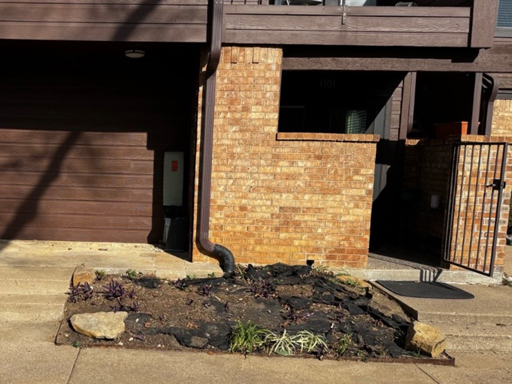 Brick townhouse facade with a curved downspout, a side gate, and a small garden bed in the foreground.