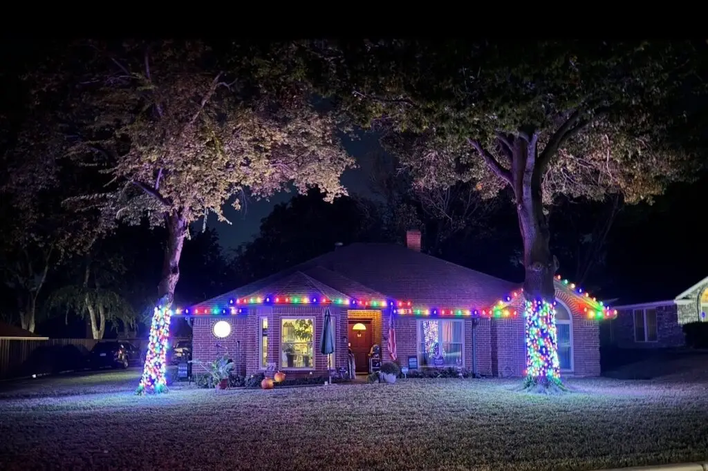 Multicolor Christmas lights along a roofline with two fully wrapped trees in the front yard