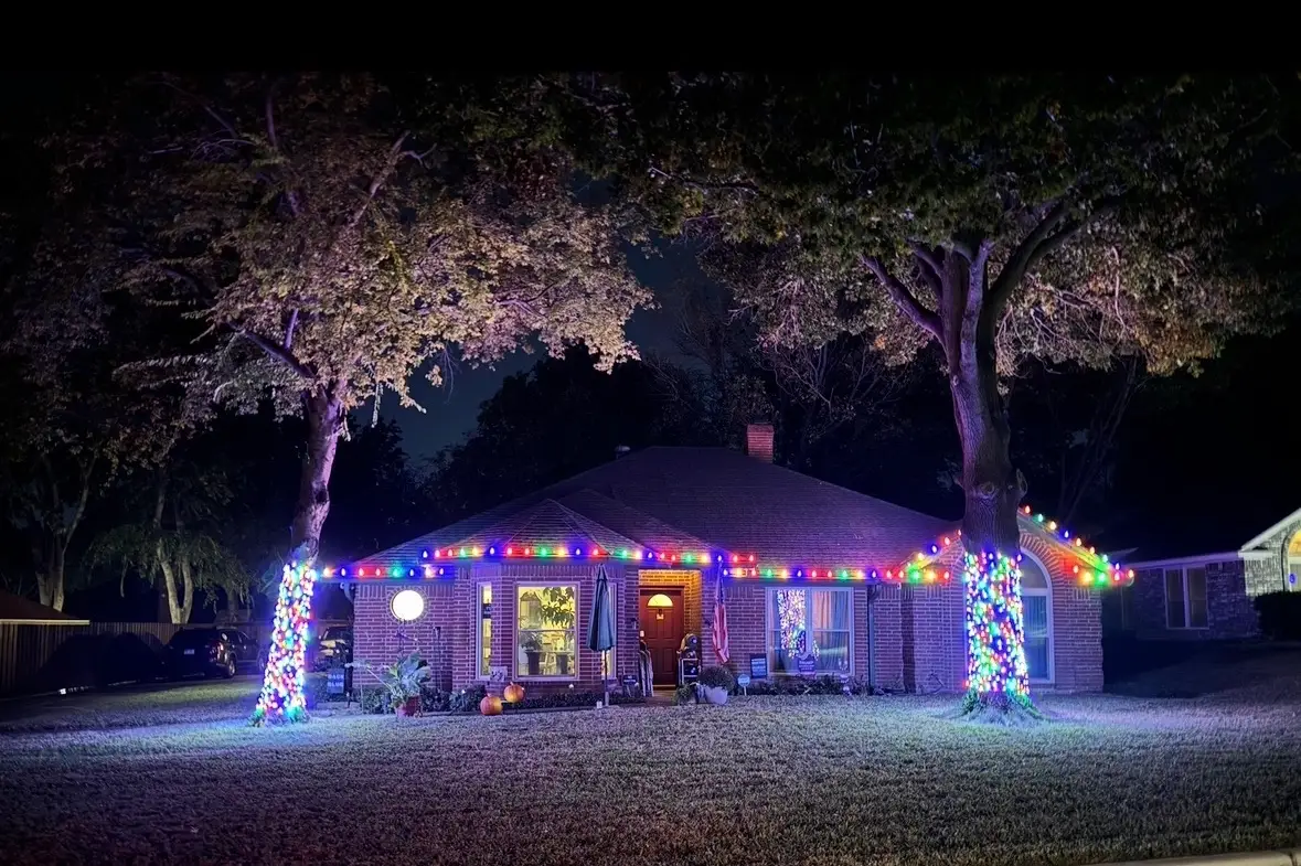 Multicolor Christmas lights along a roofline with two fully wrapped trees in the front yard