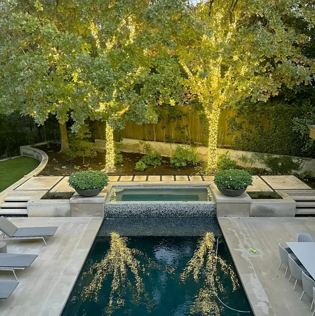 Rectangular pool with a raised spa spillway, stone coping, planted beds and boxwood planters, and lit trees along the back fence
